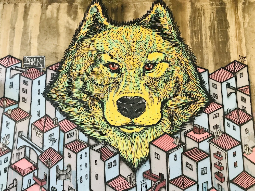 Murales di Irwin a NoLo, Milano: un lupo giallo dagli occhi rossi sovrasta una citt&agrave; stilizzata con case dai tetti rosa e dettagli urbani intricati. L&rsquo;opera di street art, vibrante e dettagliata, &egrave; un simbolo della creativit&agrave; e della cultura underground del quartiere.