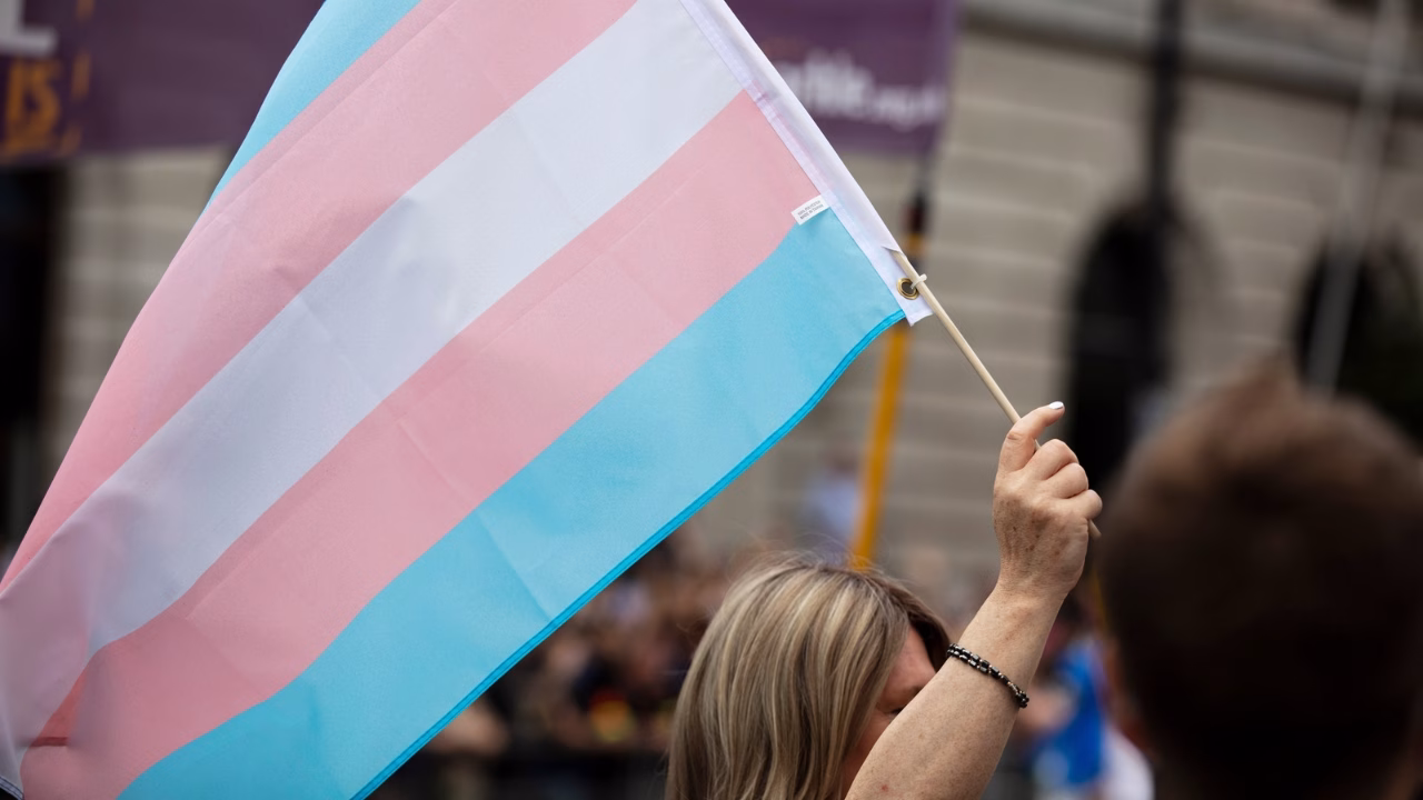 Persona con capelli biondi solleva una bandiera transgender durante una manifestazione pubblica per i diritti delle persone trans.