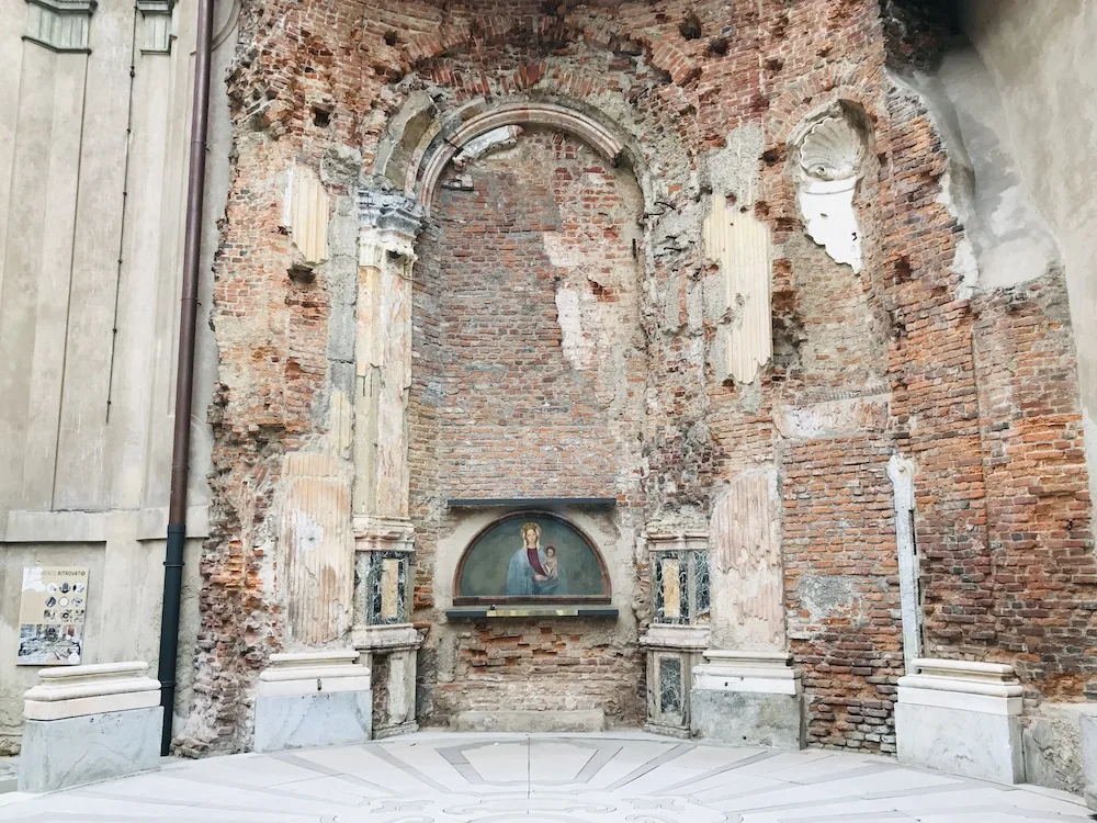 Rovine della cappella di Santa Maria alla Porta a Milano con pareti in mattoni a vista, colonne parzialmente distrutte e un&rsquo;antica nicchia con un&rsquo;icona sacra. L&rsquo;architettura porta i segni del tempo e dei bombardamenti della Seconda Guerra Mondiale, con elementi decorativi danneggiati e una pavimentazione restaurata che crea un contrasto tra il passato e il presente.