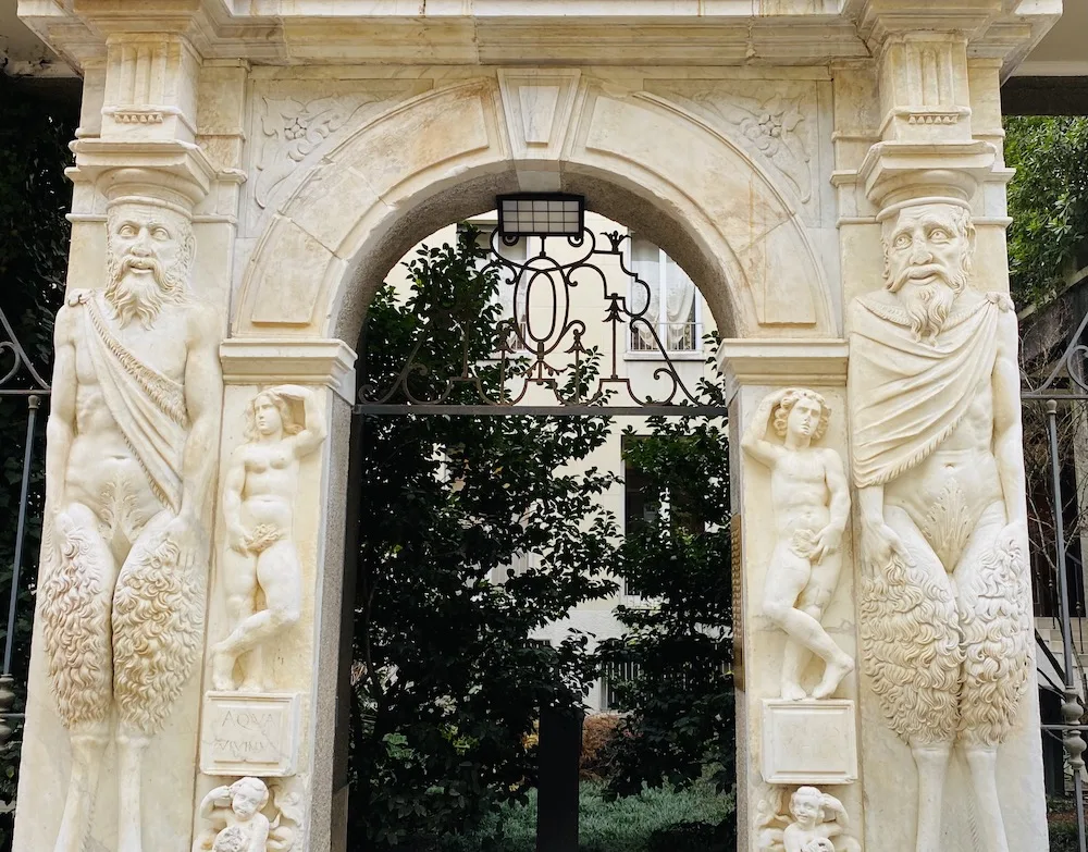Portale decorato con satiri scolpiti in pietra in via Guastalla, Milano. La struttura in marmo presenta dettagli artistici raffinati, con figure mitologiche e rilievi decorativi, incorniciando l&rsquo;ingresso di un elegante palazzo storico.