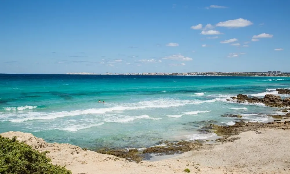 Spiaggia selvaggia con sabbia dorata e scogli a riva, bagnata dal mare turchese di Punta della Suina, Gallipoli. Sullo sfondo, la citt&agrave; di Gallipoli si affaccia sul mare cristallino del Salento, offrendo un panorama mozzafiato per chi cerca una destinazione paradisiaca in Puglia.