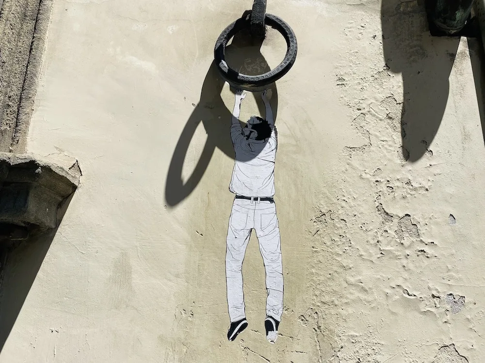 Opera di street art in Via Toscanella a Firenze, raffigurante un uomo stilizzato in bianco e nero che sembra aggrapparsi a un anello di ferro reale fissato al muro, creando un effetto tridimensionale sorprendente.