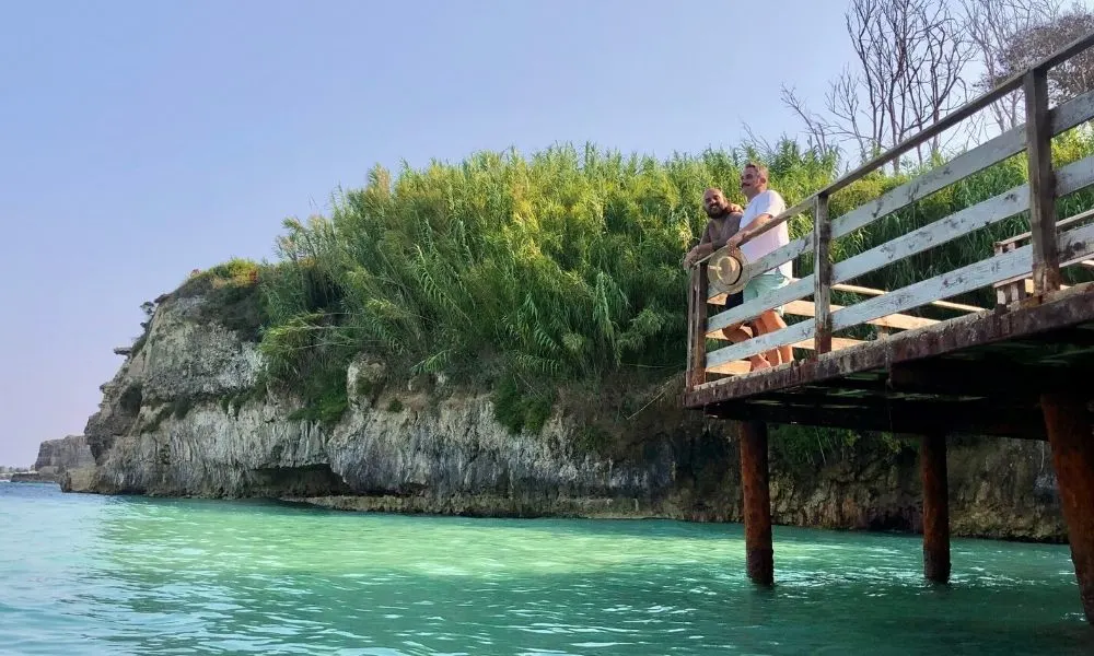 Daniele e Luigi di Gayly Planet sulla passerella di legno affacciata della Baia del Mulino d'Acqua a Otranto in Salento con un mare turchese, circondati da scogliere verdi e natura incontaminata. La scena trasmette relax e libert&agrave; in una destinazione da sogno, perfetta per una fuga romantica o un viaggio di coppia nel Salento.
