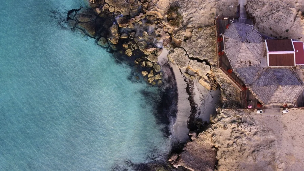 Veduta aerea di Punta della Suina, Gallipoli, con il caratteristico lido costruito su una scogliera affacciata sul mare cristallino del Salento. Le acque turchesi incontrano la costa rocciosa e una piccola spiaggia sabbiosa, creando uno scenario mozzafiato perfetto per una vacanza in Salento.