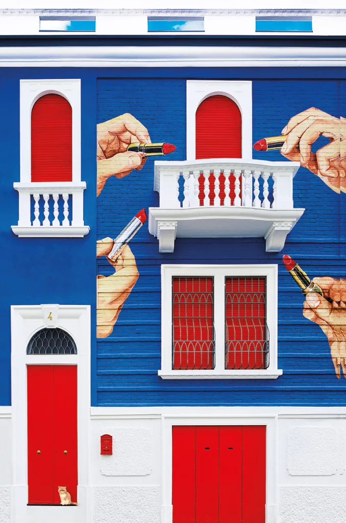 Facciata della Toiletpaper Home a Milano, caratterizzata da un murale pop-art con mani giganti che tengono rossetti rossi su sfondo blu acceso. L&rsquo;edificio presenta finestre e porte rosse con dettagli bianchi, creando un contrasto vivace e surreale tipico dello stile artistico del collettivo Toiletpaper.