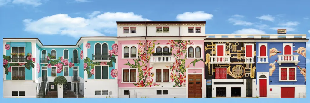 Vista panoramica della Toiletpaper Home a Milano, una serie di edifici decorati con murales artistici e surreali. Le facciate presentano motivi unici: una casa azzurra con rose rosa, una rosa con rampicanti floreali, una nera con strumenti musicali dorati e una blu con mani giganti che tengono rossetti rossi. Un&rsquo;icona dell&rsquo;arte contemporanea e della creativit&agrave; urbana nel cuore di Milano.