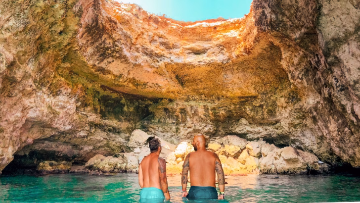 Daniele e Luigi di Gayly Planet immersi nell&rsquo;acqua cristallina esplorano la grotta sfondata, una grotta marina illuminata dal sole, circondata da imponenti pareti rocciose. Si trovano alla Baia del Mulino d'acqua a Otranto e la scena trasmette avventura e meraviglia, perfetta per chi cerca esperienze uniche nelle calette nascoste del Salento.