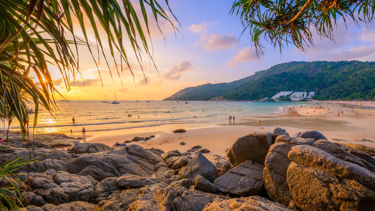 Spiaggia di Nai Harn a Phuket Thailandia al tramonto con sabbia dorata rocce in primo piano barche a vela sul mare e persone che passeggiano e fanno il bagno atmosfera tropicale e rilassante