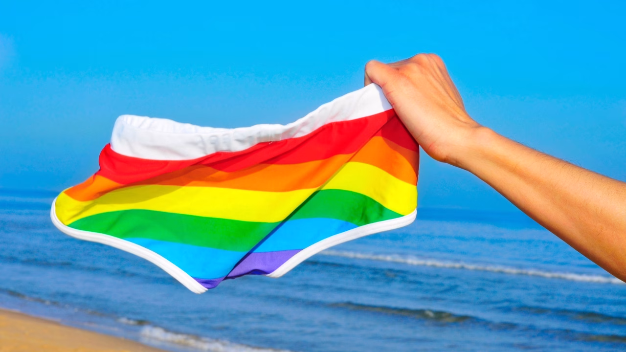 Un braccio solleva un costume da bagno a righe arcobaleno sulla spiaggia, con l&rsquo;oceano sullo sfondo e cielo azzurro limpido. L&rsquo;immagine evoca libert&agrave; e orgoglio LGBTQ+ nella cornice naturale della Platja de l&rsquo;Home Mort, iconica spiaggia gay e naturista vicino a Sitges.