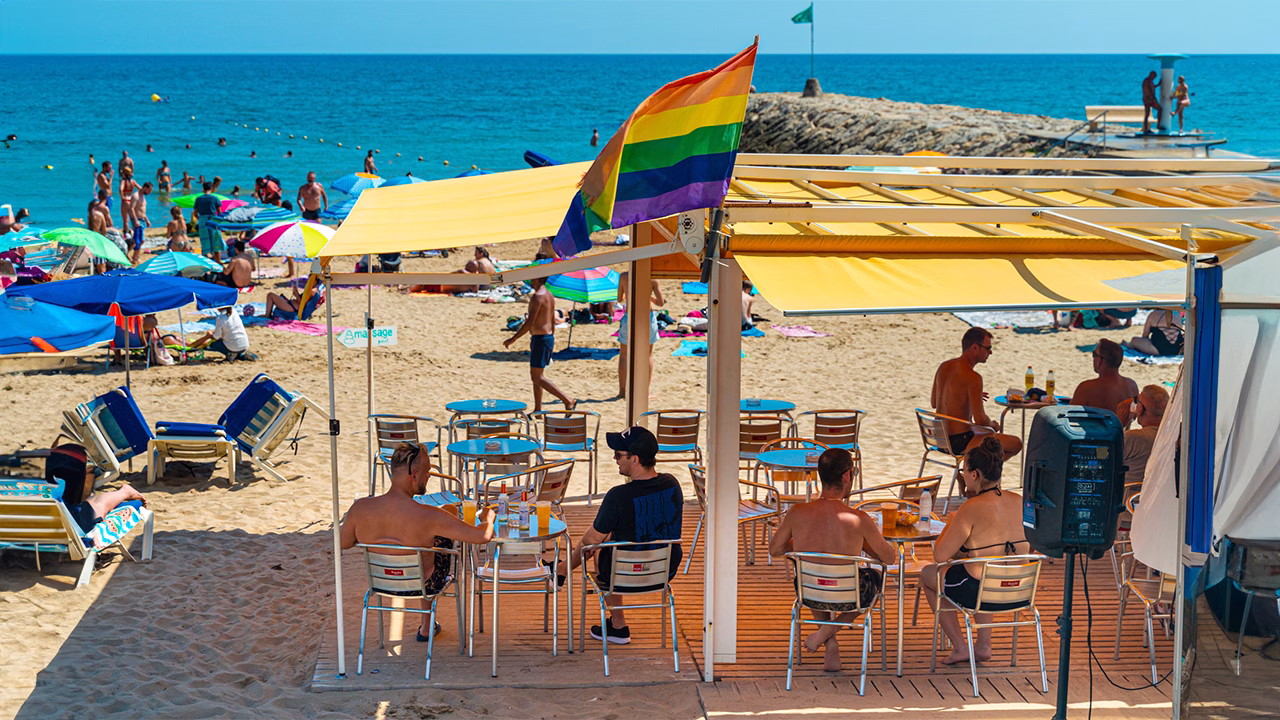 Clienti seduti sotto un gazebo giallo sorseggiano drink nel chiringuito sulla Playa de la Bassa Rodona a Sitges, una delle spiagge gay pi&ugrave; iconiche della Spagna. In alto sventola la bandiera arcobaleno, mentre sullo sfondo la spiaggia &egrave; gremita di bagnanti e ombrelloni colorati.