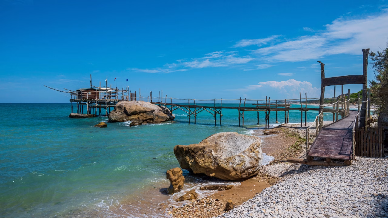 Spiaggia naturista di Lido Punta Le Morge in Abruzzo con mare turchese, grandi rocce sulla battigia e un tradizionale trabocco in legno che si estende sull&rsquo;acqua.