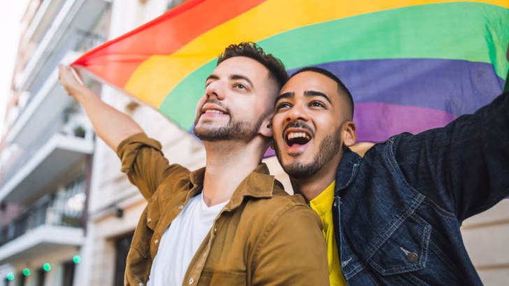 Due uomini sorridenti posano insieme all&rsquo;aperto, tenendo una grande bandiera arcobaleno sopra di loro, simbolo dell&rsquo;orgoglio LGBTQ+. Uno indossa una camicia marrone e l&rsquo;altro una giacca di jeans con una maglia gialla, entrambi con un&rsquo;espressione di gioia e libert&agrave;. Sullo sfondo si vedono edifici urbani, creando un&rsquo;atmosfera di celebrazione e inclusivit&agrave;.