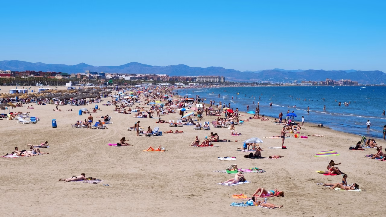 Spiaggia affollata di Playa Malvarrosa a Valencia, con persone che prendono il sole, si rilassano su lettini e fanno il bagno nel mare calmo. Sullo sfondo, palme, ombrelloni di paglia e la citt&agrave; che si estende lungo la costa, perfetta per una giornata LGBTQ+ friendly.