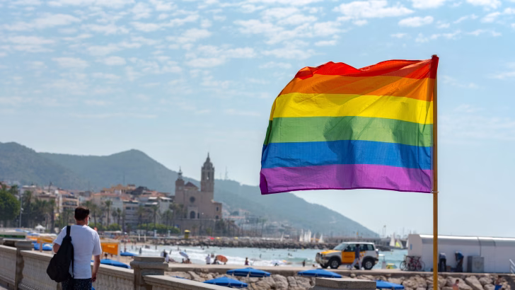 Una grande bandiera arcobaleno sventola sul lungomare di Sitges, con vista sulla spiaggia e sul centro storico dominato dalla chiesa di San Bartolomeo e Santa Tecla. L&rsquo;immagine celebra l&rsquo;atmosfera inclusiva e le famose spiagge gay e nudiste di Sitges, meta LGBTQ+ tra le pi&ugrave; iconiche della Spagna.