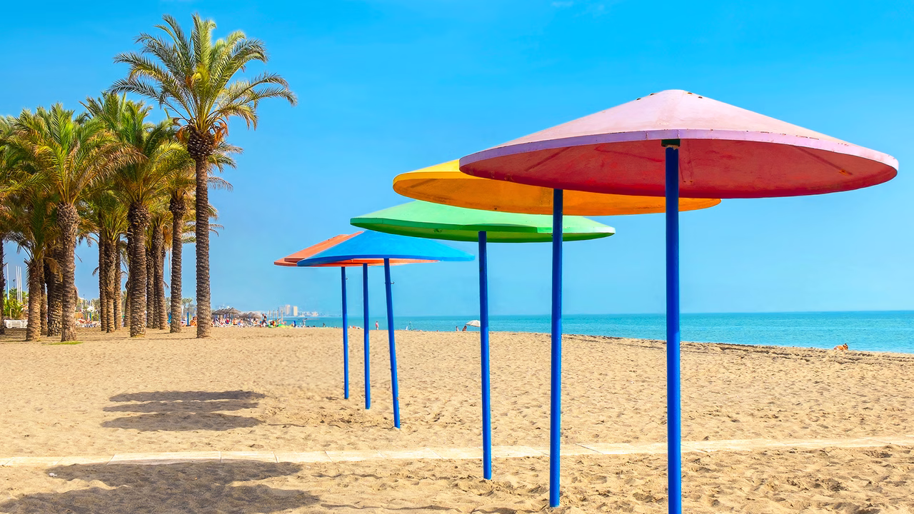 Spiaggia gay a Torremolinos con file di ombrelloni colorati in stile arcobaleno che richiamano la bandiera LGBTQ+, affiancati da alte palme. Un&rsquo;icona dell&rsquo;ospitalit&agrave; gay-friendly della Costa del Sol, perfetta per una vacanza inclusiva tra sole e mare.