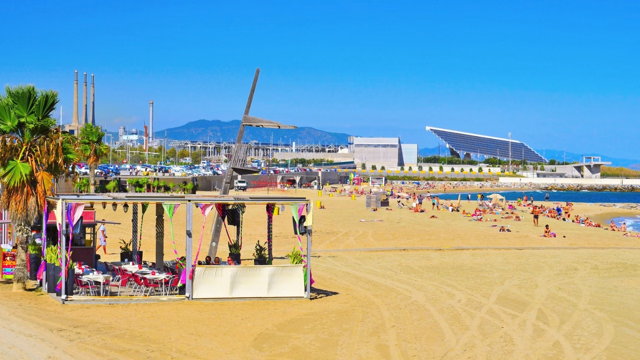 Spiaggia gay e nudista di Barcellona Mar Bella, con chiringuito decorato in stile vivace in primo piano e numerosi bagnanti distesi al sole. Sullo sfondo si intravede il grande pannello solare fotovoltaico del Parc del F&ograve;rum, simbolo architettonico della zona.