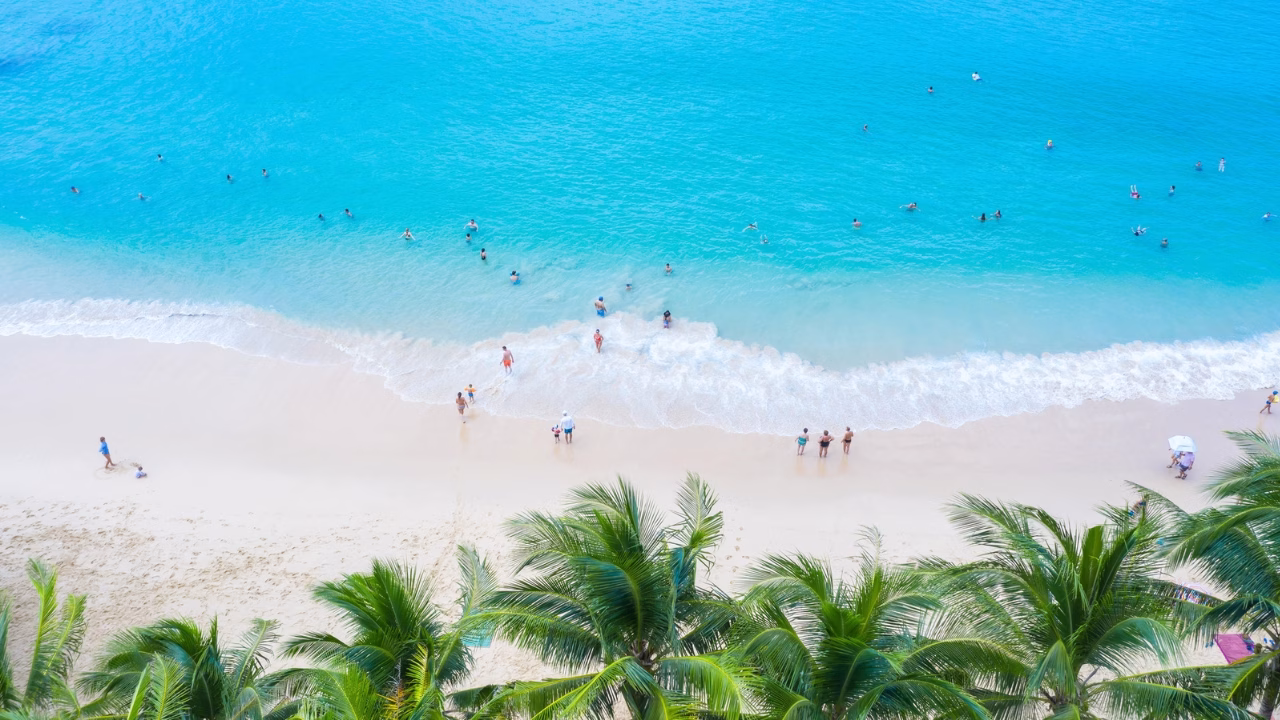Vista dall&rsquo;alto della spiaggia gay friendly tropicale di Surin Beach a Phuket con palme in primo piano, sabbia bianca e mare turchese, dove bagnanti nuotano e si rilassano vicino alla riva. L&rsquo;atmosfera &egrave; soleggiata e vivace, tipica di una meta balneare esotica.