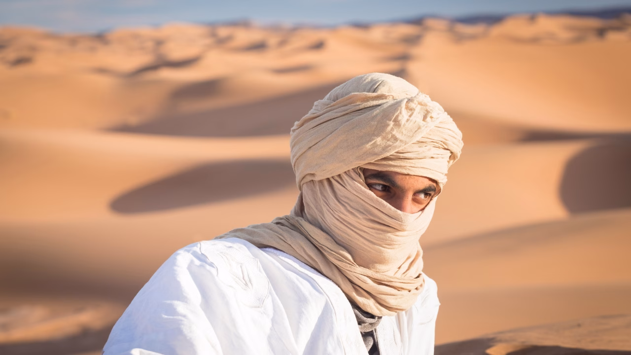 Uomo con turbante beige e tunica bianca nel deserto, con lo sguardo rivolto verso l&rsquo;orizzonte tra le dune dorate. Outfit tradizionale per proteggersi dal sole e dal vento, ideale per chi cerca consigli su come vestirsi nel deserto. Viaggio nel deserto, abbigliamento per il deserto, cosa indossare nel deserto.