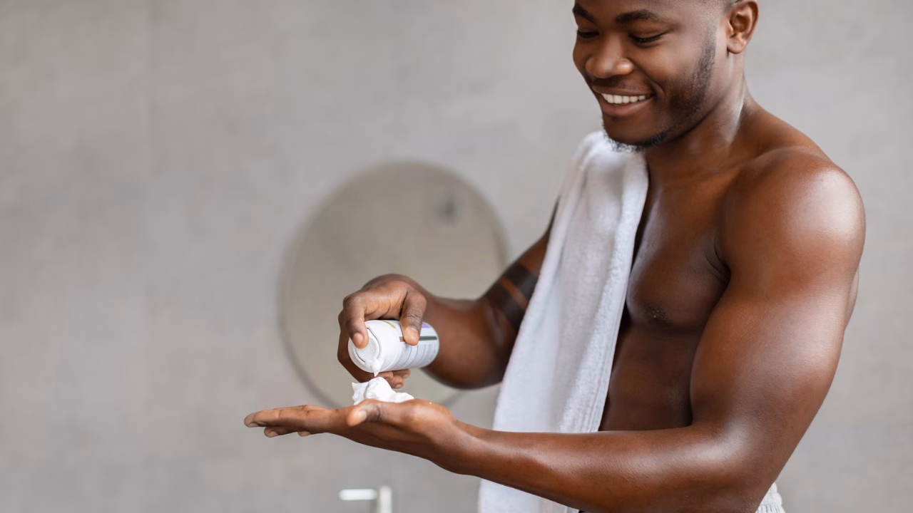 Un uomo sorridente, con un asciugamano bianco sulle spalle, versa della crema da barba sulla mano mentre si prepara davanti allo specchio in un bagno minimalista. L&rsquo;immagine trasmette un senso di benessere e cura personale.