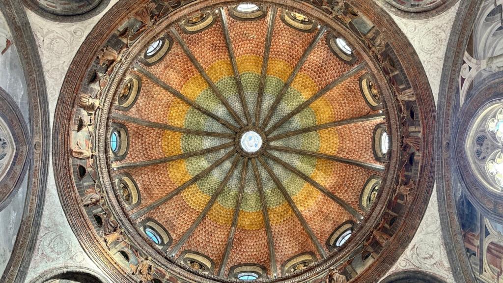 La cupola affrescata della Cappella Portinari nella Basilica di Sant&rsquo;Eustorgio a Milano, conosciuta per il suo particolare effetto cromatico ad arcobaleno. La decorazione geometrica e radiale crea un suggestivo gioco di colori dal verde al giallo e al rosso, illuminato dalle finestre circolari che ne esaltano la bellezza artistica e architettonica.