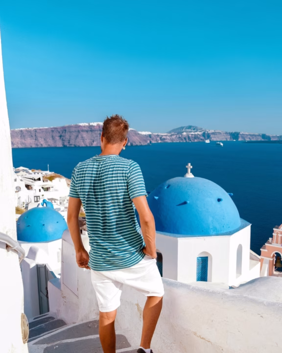 Uomo di spalle con maglietta a righe e pantaloncini bianchi che osserva i caratteristici edifici bianchi con cupole blu di Santorini, Grecia, con vista sul mare azzurro e le scogliere vulcaniche in lontananza.