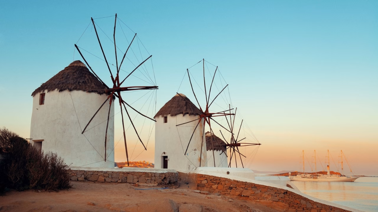 Antichi mulini a vento bianchi con tetti di paglia a Mykonos, Grecia, illuminati dalla luce calda del tramonto, con il mare calmo e alcune barche a vela sullo sfondo.