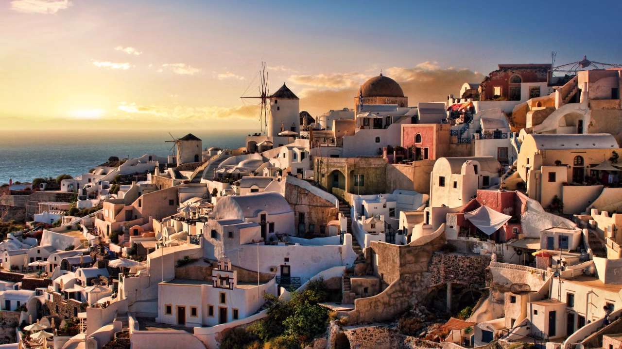 Panoramica di Oia a Santorini al tramonto, con case bianche e color pastello costruite sulle scogliere e un mulino a vento tradizionale che si staglia contro il cielo dorato.