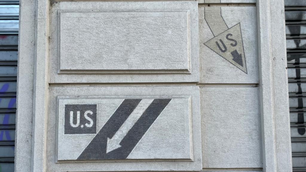 Simboli di guerra incisi sulla facciata di un palazzo in Piazzale di Porta Lodovica a Milano, risalenti alla Seconda Guerra Mondiale. Questi segni, con la scritta &lsquo;U.S.&rsquo; e frecce direzionali, indicavano la presenza di rifugi antiaerei o percorsi strategici per le truppe alleate.