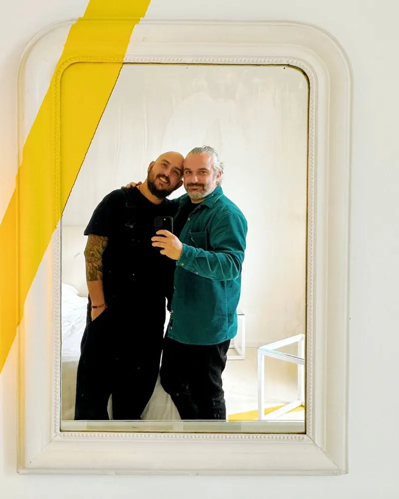 Daniele e Lugii di Gayly Planet in una camera da letto moderna e minimalista con arredi total white, letto matrimoniale al centro, pareti bianche decorate da una linea gialla continua e due poltrone tonde vicino a una finestra luminosa. Questo &egrave; il Le Windsor Jungle Art Hotel, un hotel gay friendly a Nizza con un'atmosfera rilassante e design contemporaneo