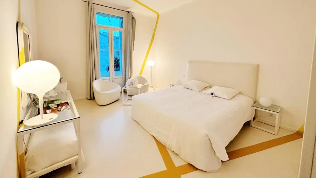 Camera da letto moderna e minimalista con arredi total white, letto matrimoniale al centro, pareti bianche decorate da una linea gialla continua e due poltrone tonde vicino a una finestra luminosa. Questo &egrave; il Le Windsor Jungle Art Hotel, un hotel gay friendly a Nizza con un'atmosfera rilassante e design contemporaneo