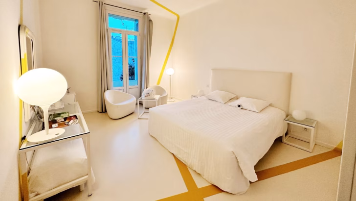 Camera da letto moderna e minimalista con arredi total white, letto matrimoniale al centro, pareti bianche decorate da una linea gialla continua e due poltrone tonde vicino a una finestra luminosa. Questo &egrave; il Le Windsor Jungle Art Hotel, un hotel gay friendly a Nizza con un'atmosfera rilassante e design contemporaneo