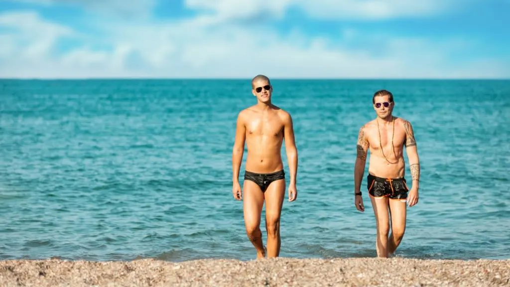 Due ragazzi gay escono sorridenti dal mare sulla spiaggia gay della Lecciona in Versilia, con l’acqua cristallina e il cielo azzurro sullo sfondo.