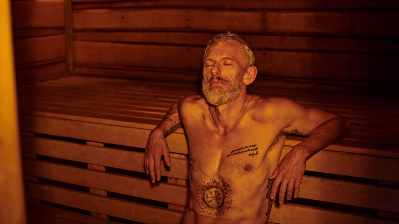 Un uomo maturo con barba grigia e tatuaggi sul petto e sulle braccia &egrave; seduto rilassato in una sauna gay di Torino. Ha gli occhi chiusi e un&rsquo;espressione di calma, mentre la luce calda illumina il suo viso e il suo corpo. L&rsquo;atmosfera &egrave; intima e avvolgente, enfatizzando il senso di benessere e relax.