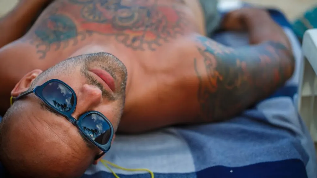Uomo tatuato sdraiato su un telo da mare blu e bianco, con occhiali da sole riflettenti palme, mentre si rilassa su una spiaggia gay in Puglia