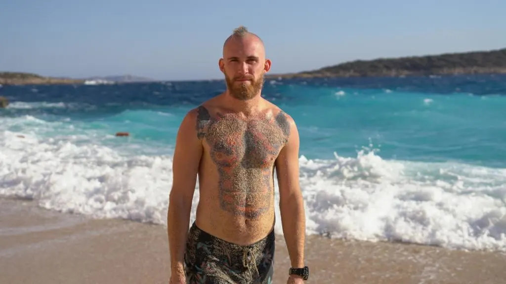 Ragazzo gay con barba e tatuaggi in costume da bagno, coperto di sabbia, posa su una spiaggia toscana con mare mosso e acque turchesi sullo sfondo.