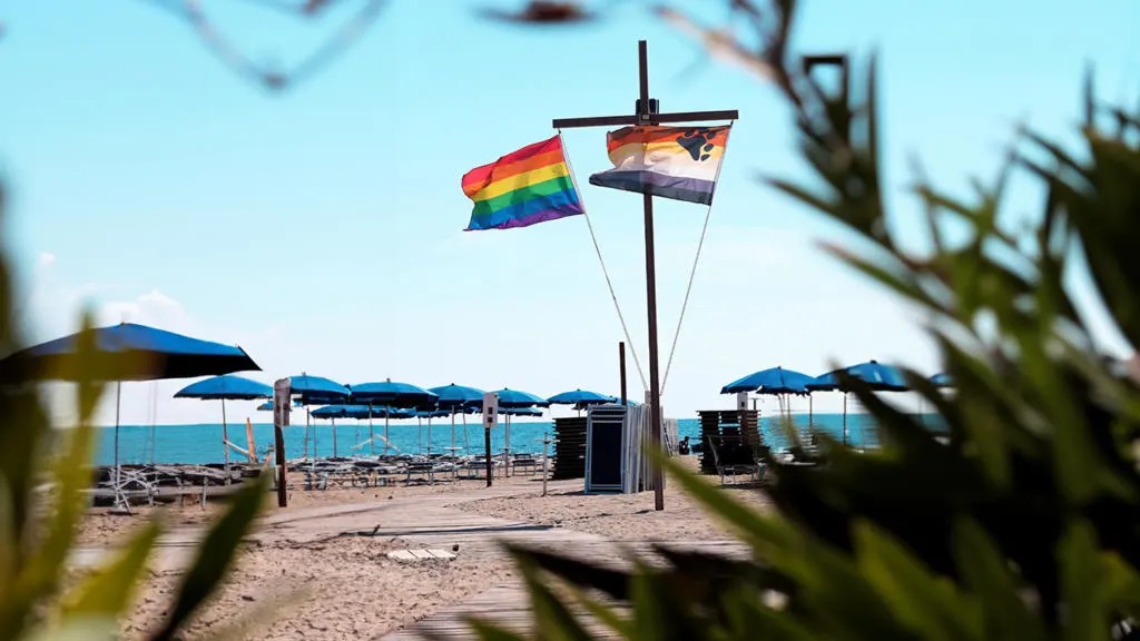 Vista sulla spiaggia gay del Mama Beach a Torre del Lago, con ombrelloni blu ordinati lungo la riva e bandiere arcobaleno e bear pride che sventolano al vento sotto un cielo limpido.
