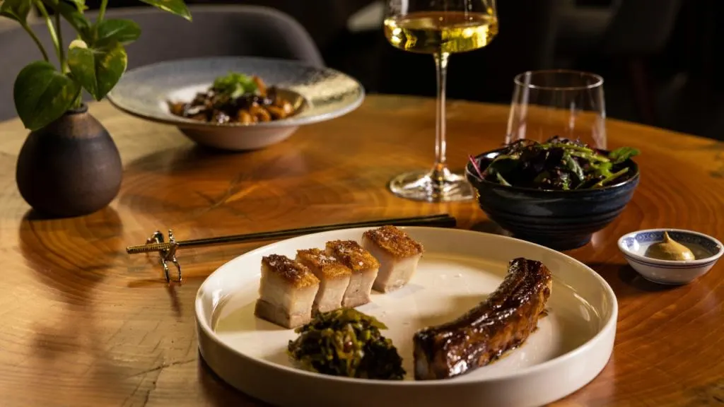 Piatto di maiale croccante e costine glassate servito al ristorante &lsquo;MU dimsum&rsquo; di Milano, accompagnato da verdure fermentate e insalata. L&rsquo;elegante presentazione su un tavolo in legno con bacchette dorate e un calice di vino bianco esalta l&rsquo;atmosfera raffinata del locale specializzato in cucina cantonese.