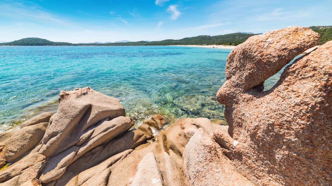 Vista mozzafiato dalla spiaggia di Liscia Ruja in Costa Smeralda, con grandi rocce di granito modellate dal vento in primo piano, mare cristallino dalle sfumature turchesi e fondale visibile, circondato da una costa verde e incontaminata.
