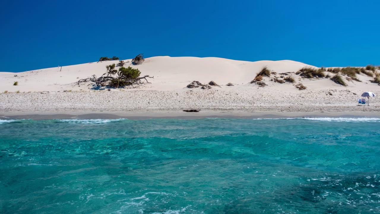 Spiaggia gay in Sardegna di Is Arenas con mare turchese e limpido in primo piano, seguita da sabbia chiara e dune ondulate punteggiate da piccoli arbusti sotto un cielo azzurro intenso.