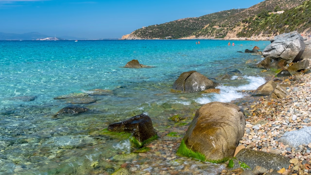 Scorcio della spiaggia gay in Sardegna di Mari Pintau in Sardegna, con acque cristalline che sfumano dal turchese al blu intenso, ciottoli levigati e grandi rocce disseminate lungo la riva, immersa in un contesto naturale incontaminato.