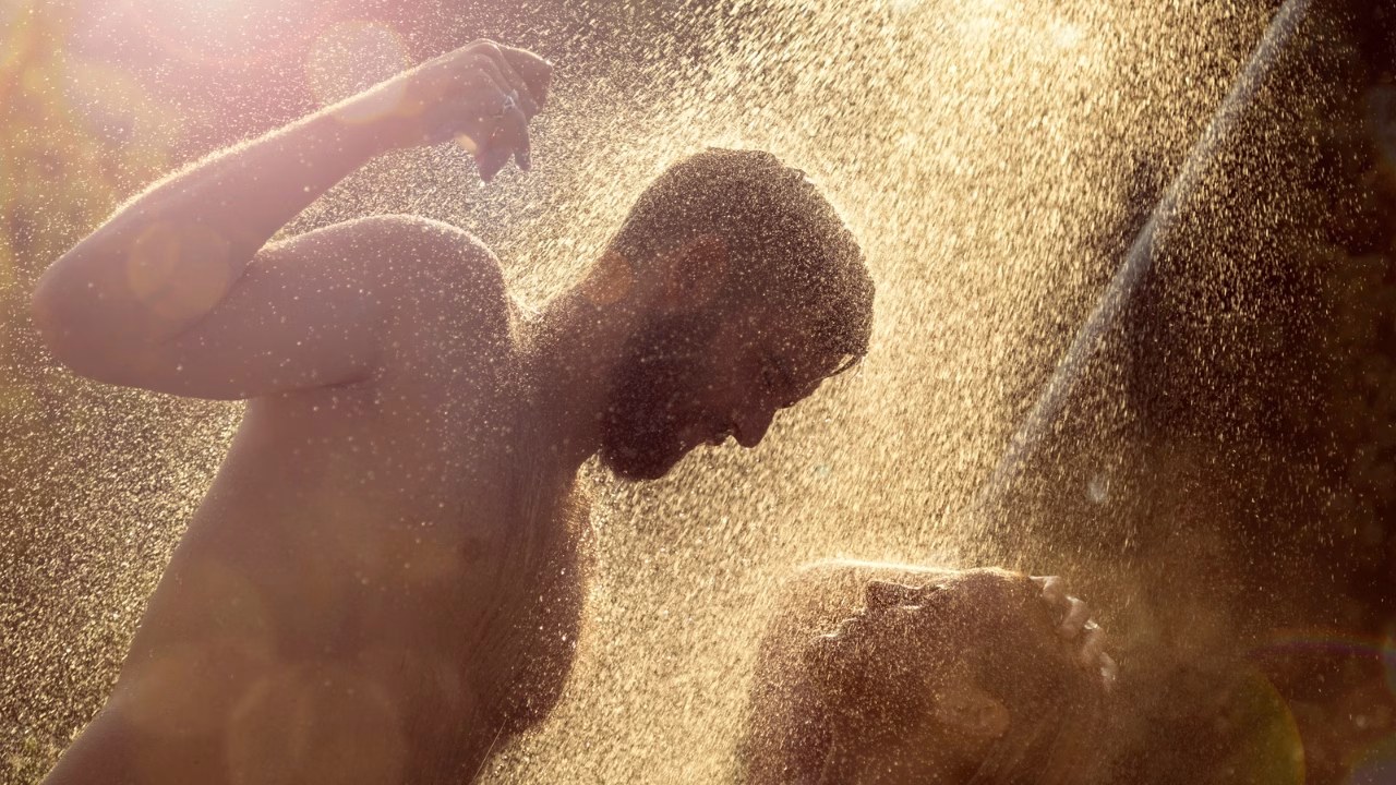 Due uomini sotto una doccia esterna in controluce, avvolti da gocce d&rsquo;acqua dorate illuminate dal sole. La scena trasmette intimit&agrave;, spensieratezza e una forte connessione in un momento di relax all&rsquo;aperto.