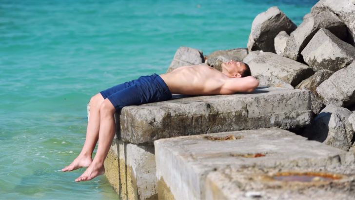 Ragazzo sdraiato al sole su un molo di cemento in riva al mare, con acqua turchese sullo sfondo e blocchi di roccia attorno. Una scena tipica delle spiagge gay e naturiste della Liguria, ideali per relax e incontri.