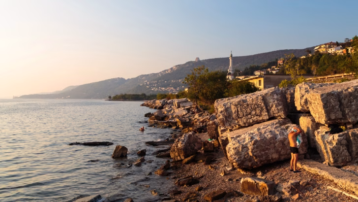 Tratto roccioso di costa vicino a Trieste al tramonto, con una persona in costume che si asciuga vicino a grandi blocchi di pietra, mentre alcune persone fanno il bagno in mare e sullo sfondo si intravede il Faro della Vittoria