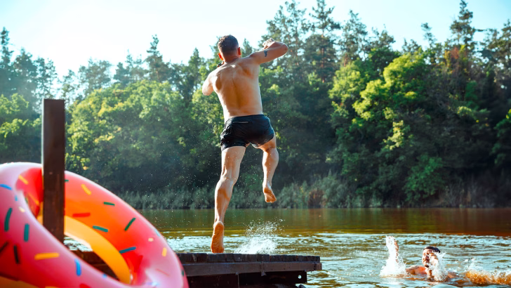 Due uomini si divertono in un lago circondato dalla natura: uno salta dal pontile mentre l&rsquo;altro lo aspetta gi&agrave; in acqua. A sinistra, un grande salvagente colorato a forma di ciambella aggiunge un tocco giocoso alla scena estiva