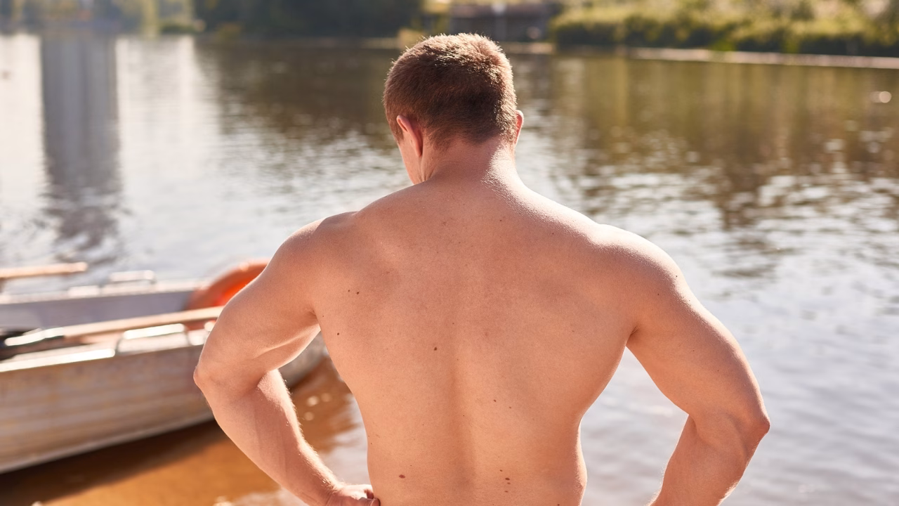 Uomo muscoloso e a torso nudo in piedi sulla riva di un lago alpino, con le mani sui fianchi e lo sguardo rivolto verso l&rsquo;acqua calma. L&rsquo;atmosfera rilassata richiama le spiagge gay e nudiste della Valle d&rsquo;Aosta immerse nella natura.