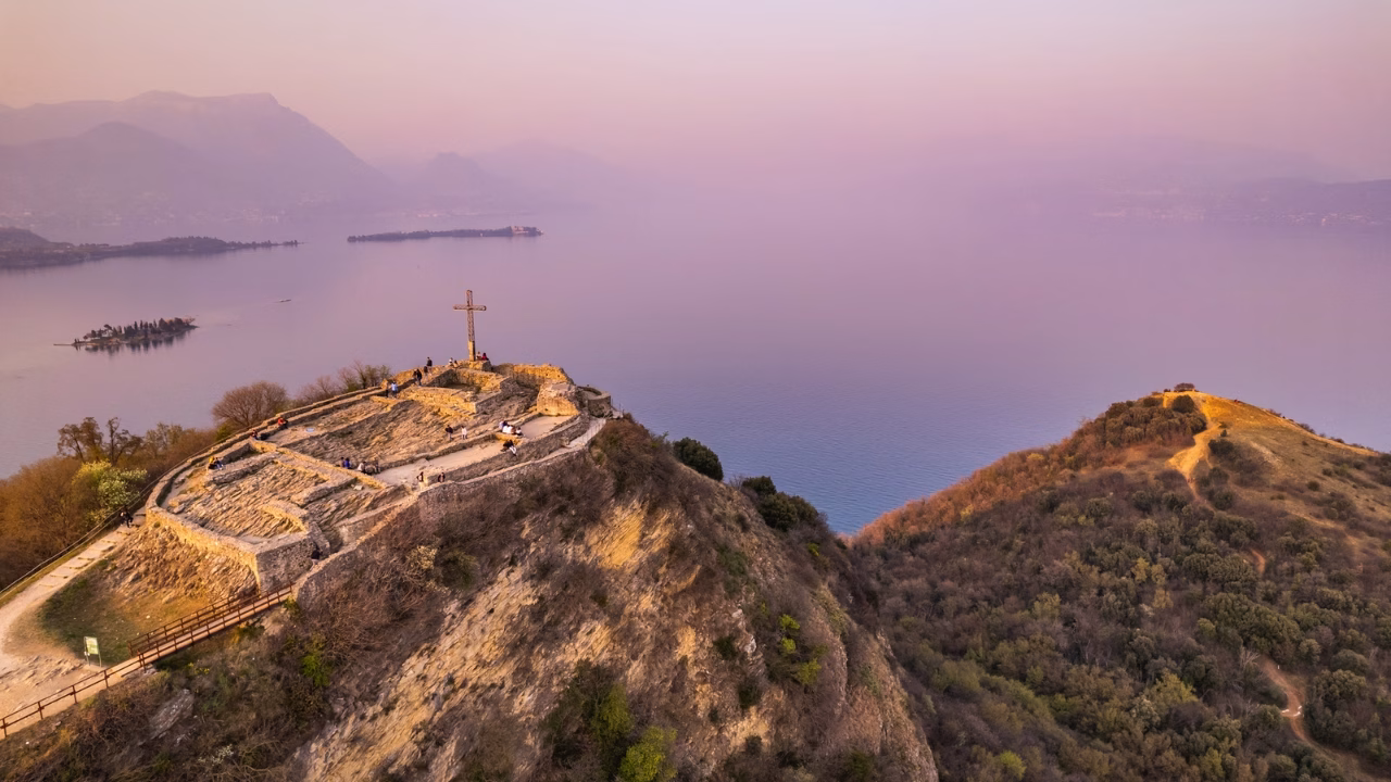 Veduta aerea della Rocca di Manerba al tramonto, con le rovine dell&rsquo;antica fortificazione e una grande croce panoramica che si affaccia sul Lago di Garda. Il paesaggio tranquillo e suggestivo &egrave; circondato da colline boscose e acque placide.
