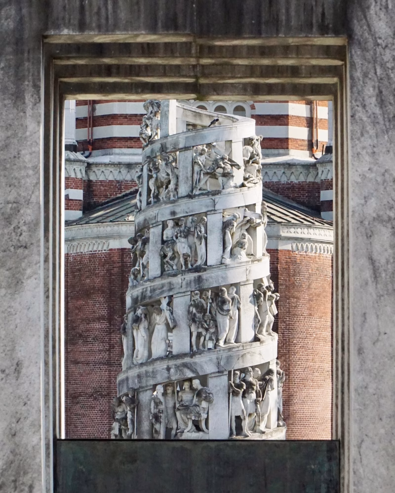 Dettaglio della suggestiva scultura a spirale del Cimitero Monumentale di Milano, incorniciata da un&rsquo;apertura architettonica. L&rsquo;opera, ricca di figure scolpite, si staglia sullo sfondo della Cappella Centrale, uno dei simboli di Milano segreta e insolita
