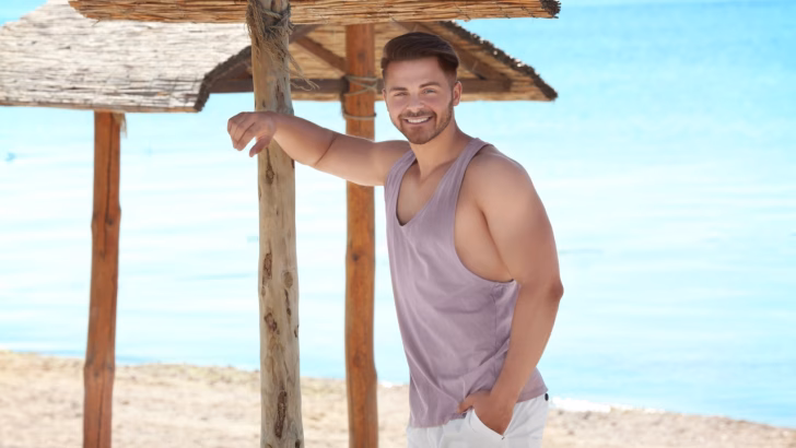 Ragazzo sorride sotto un ombrellone di paglia in una delle spiagge gay e naturiste della Campania, una delle destinazioni LGBTQ+ pi&ugrave; rilassanti della regione, perfetta per chi cerca libert&agrave;, mare e inclusivit&agrave;.