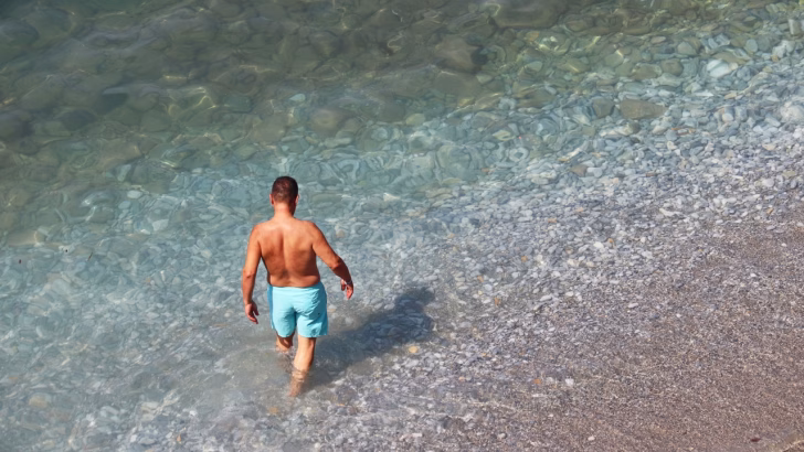 Uomo di spalle che entra nelle acque cristalline di una spiaggia di ciottoli, simbolo della tranquillit&agrave; e del turismo inclusivo delle spiagge gay nelle Marche.