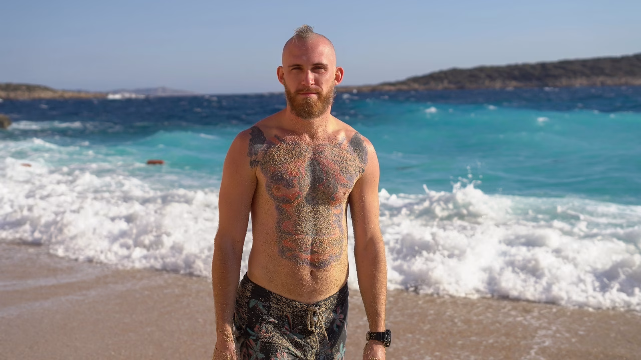 Uomo con mohawk e tatuaggi su petto e braccia, in piedi su una spiaggia sabbiosa con il torso coperto di sabbia, onde che si infrangono sullo sfondo e mare turchese agitato